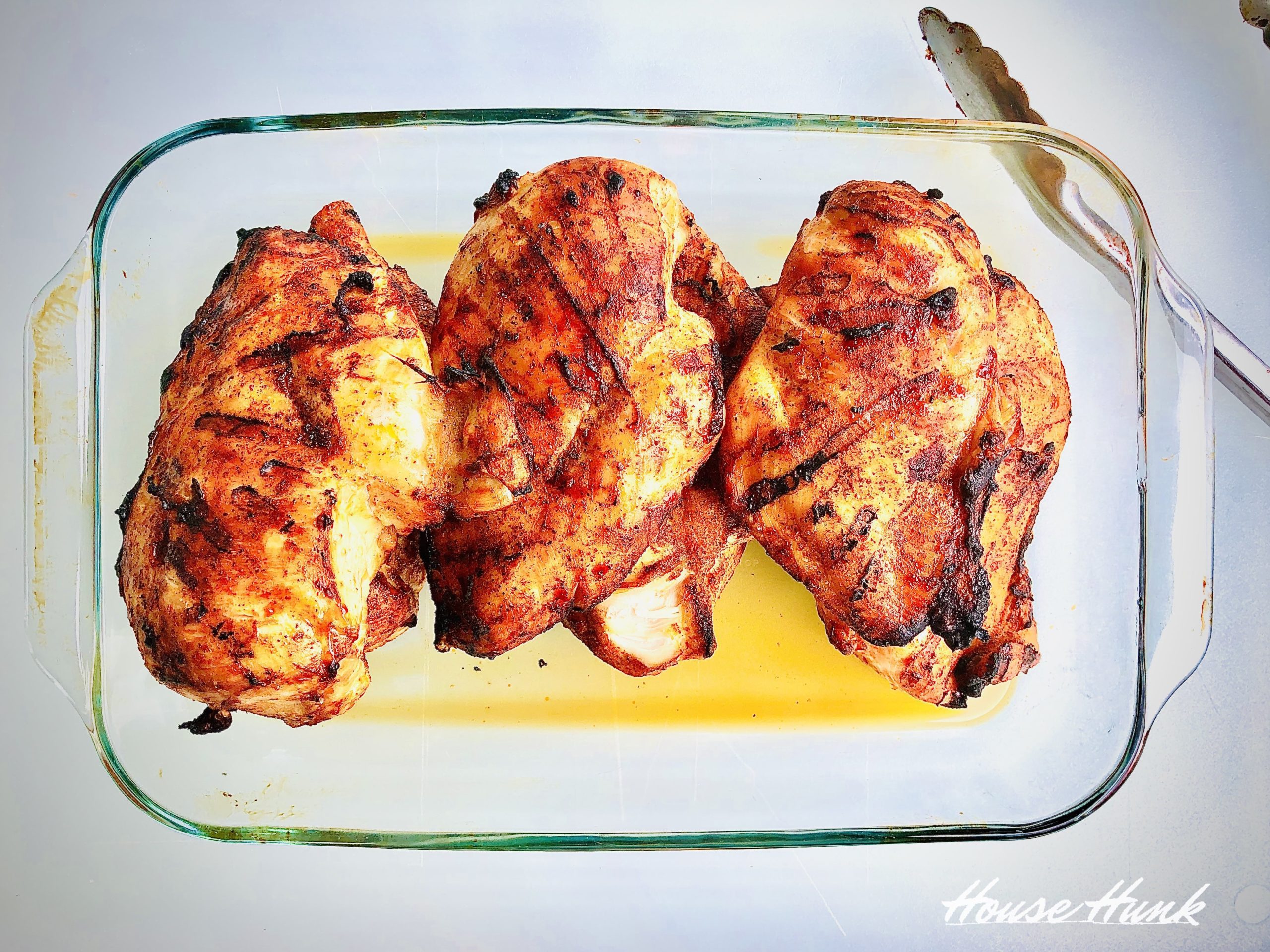 Three grilled chicken breasts in a glass baking dish with a fork and a watermark on a white background