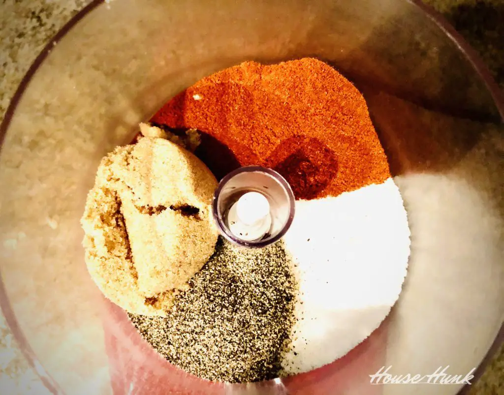 A food processor bowl with brown sugar, paprika, salt, and black pepper arranged in a circle around a metal blade on a dark countertop.