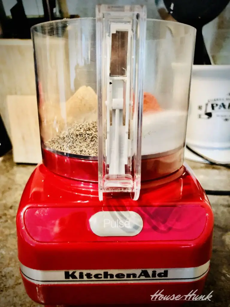 A red KitchenAid food processor filled with ground spices on a kitchen counter. The food processor has a clear plastic bowl with a white lid and a white pusher. There is a white mug with black text and a wooden knife block in the background.