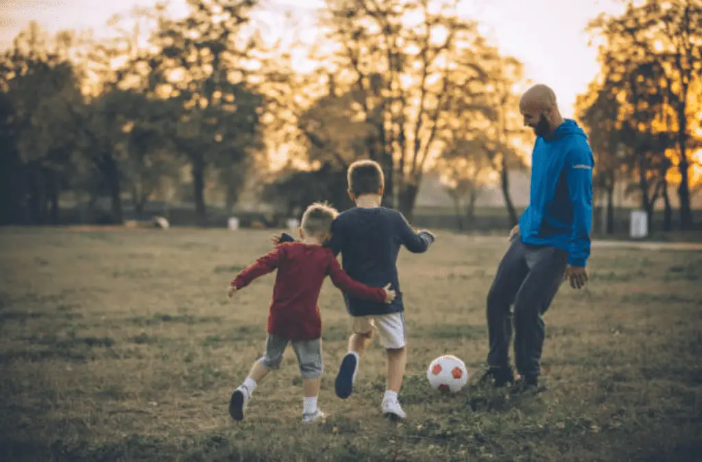 A photo of a father and two sons playing soccer in a park at sunset, wearing blue and red jackets.