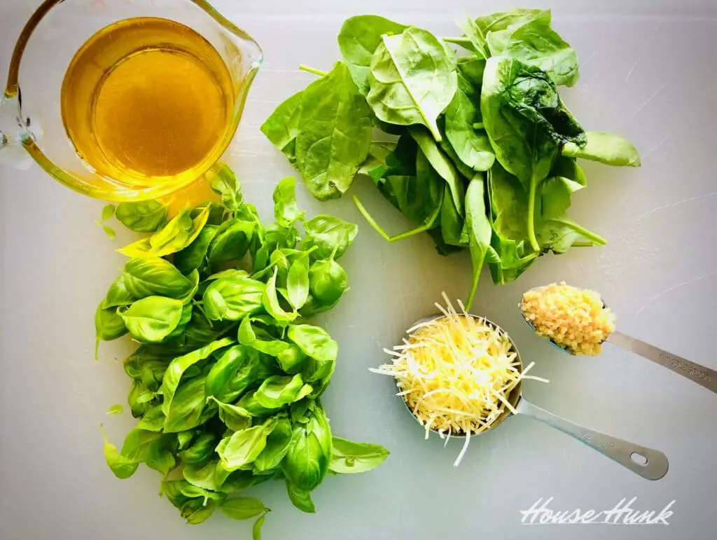An overhead photo of garlic, parmesan cheese, olive oil, spinach leaves, and basil leaves on a white cutting board.