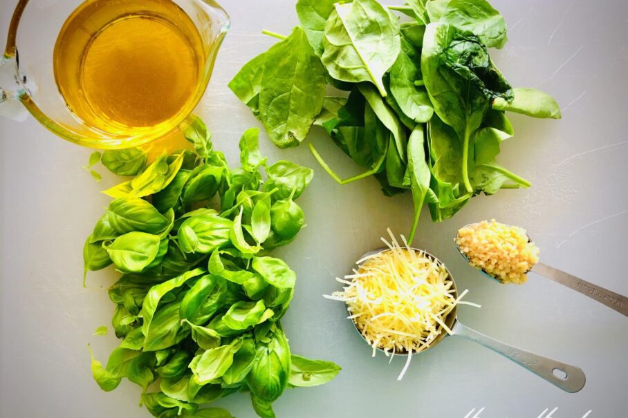An overhead photo of garlic, parmesan cheese, olive oil, spinach leaves, and basil leaves on a white cutting board.