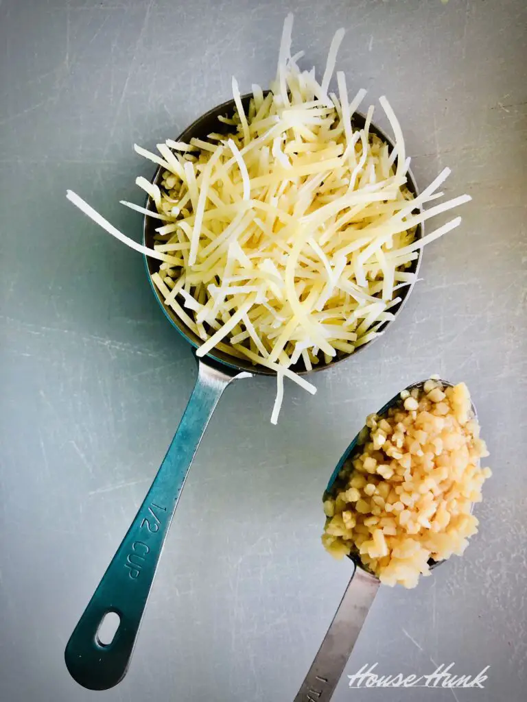 A measuring cup with shredded parmesan cheese next to a tablespoon of minced garlic to be used in making nut-free basil pesto.