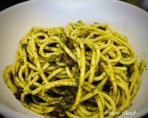 A close-up photo of noodles with nut-free pesto sauce in a white bowl with a dark background.