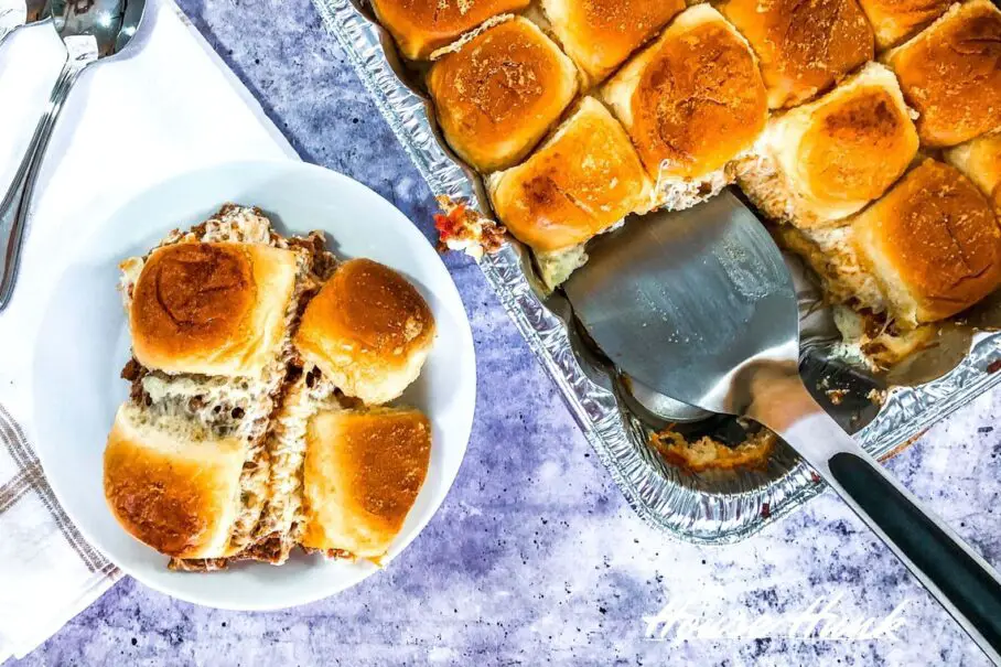 An overhead image of a tray of lasagna sliders being served out of an aluminum lasagna tray onto a white plate with a spatula.