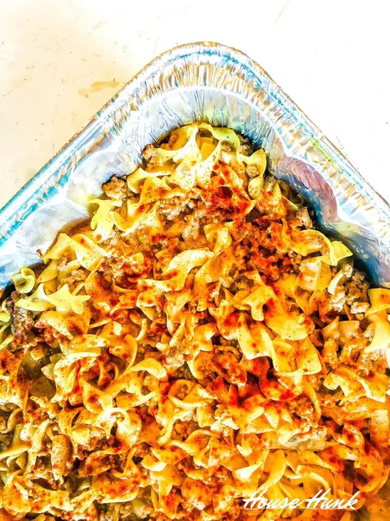 A tray of amish country casserole in an aluminum foil tray. The pasta is a mix of different shapes and sizes, with a layer of paprika on top. The dish has a tomato-based sauce with chunks of meat and vegetables mixed in. The background is a white surface with some scuff marks and crumbs. The text “HouseHunk” is in the bottom right corner of the image.