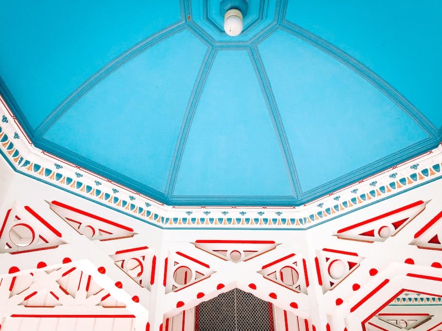 A photo of the blue ceiling and white walls of an octagonal dome with a light and red patterns.