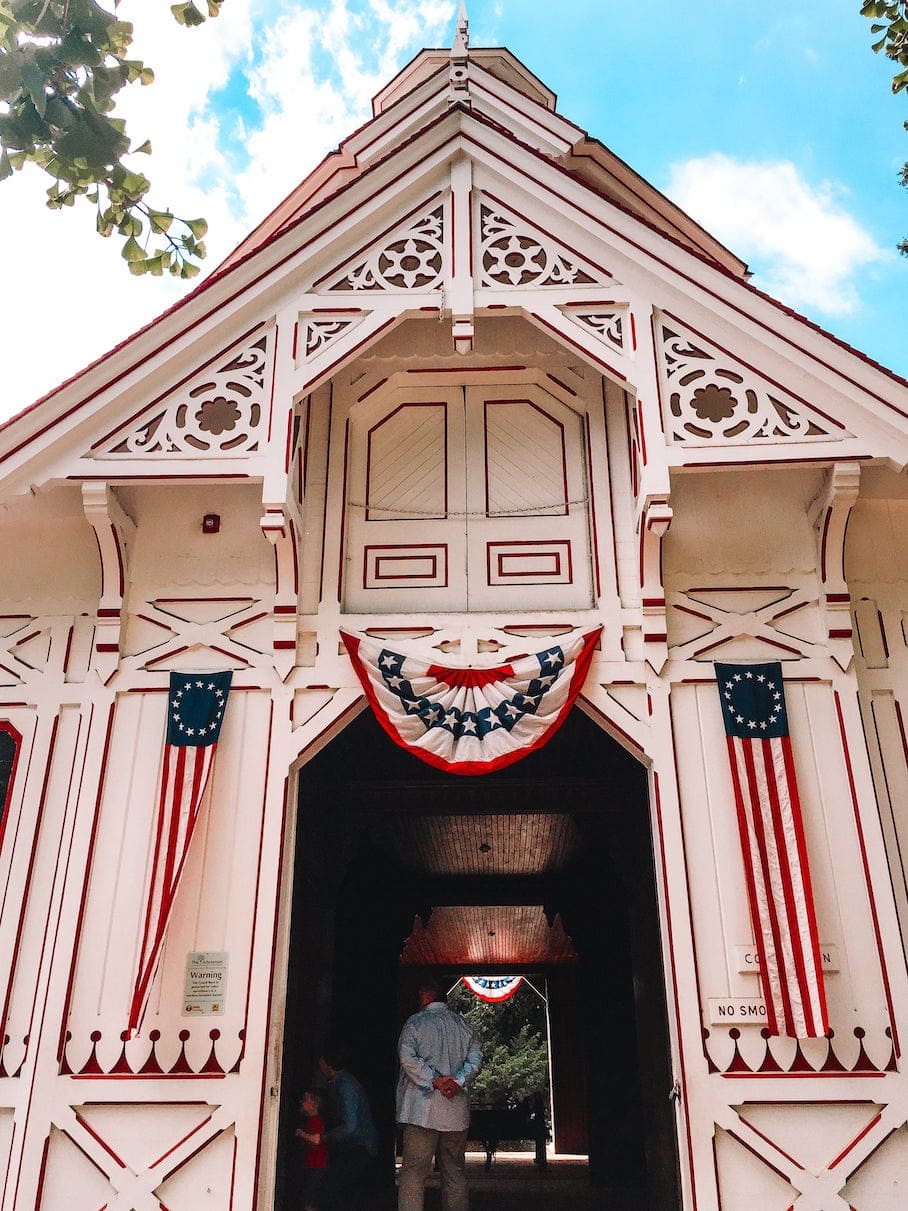 A photo of a white wooden stable building with American flags and a man inside, against a blue sky with trees.