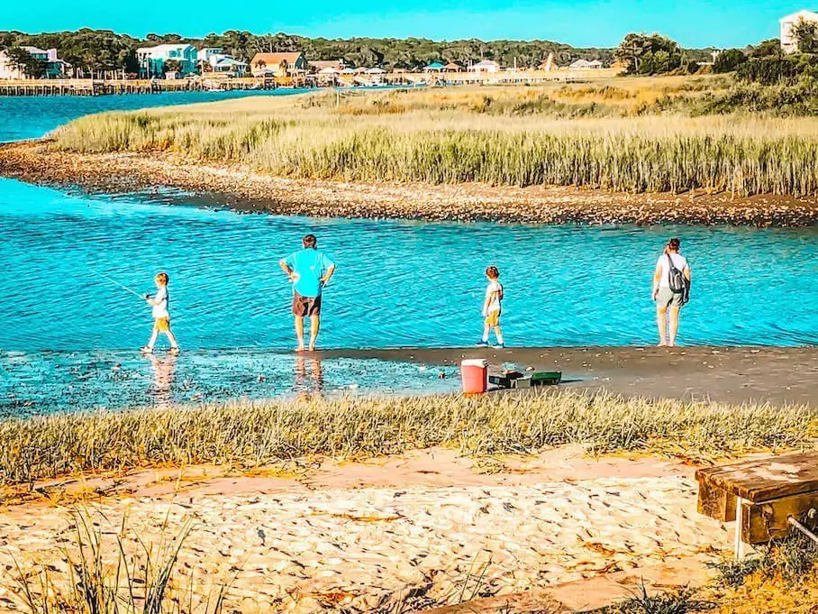 A photo of a group of people fishing on a sandy beach with tall grasses. They are holding fishing rods and looking at the calm blue water. There is a red cooler and a green bucket on the beach. The background shows a wooden pier and buildings in the distance. The photo has a blue-green filter applied to it.