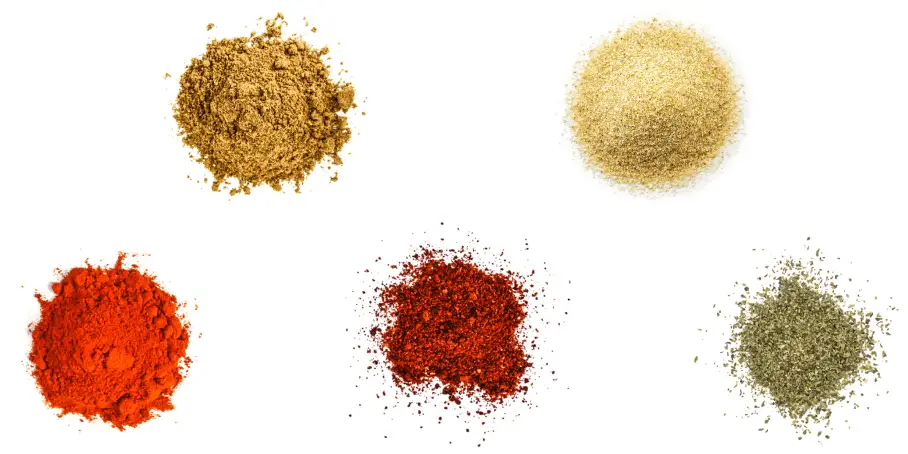 A photorealistic image of ingredients for Homemade Chili Powder consisting of five piles of different spices and herbs on a white background.