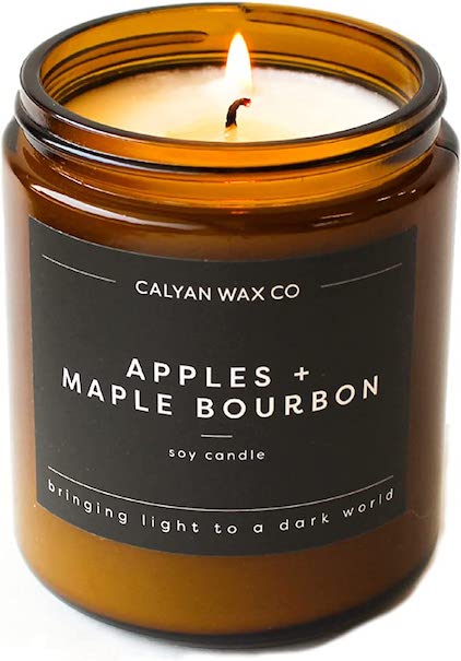A brown glass jar of Calyan Wax Co. soy candle with apples and maple bourbon scent and a black lid, lit on a white background.