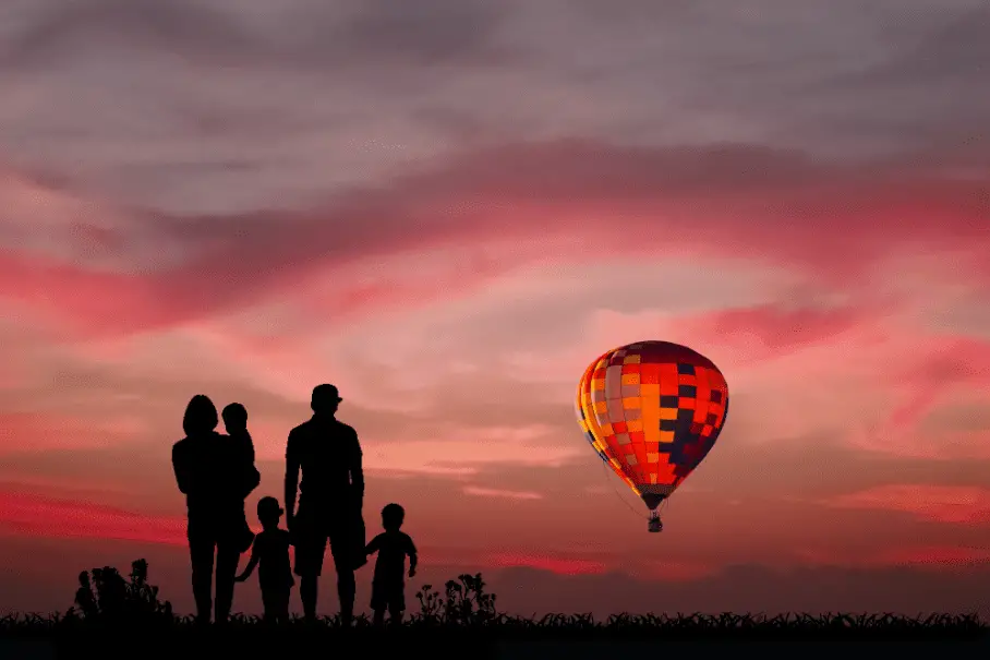 A family of five silhouetted on a hill with a sunset sky and a hot air balloon in the background.