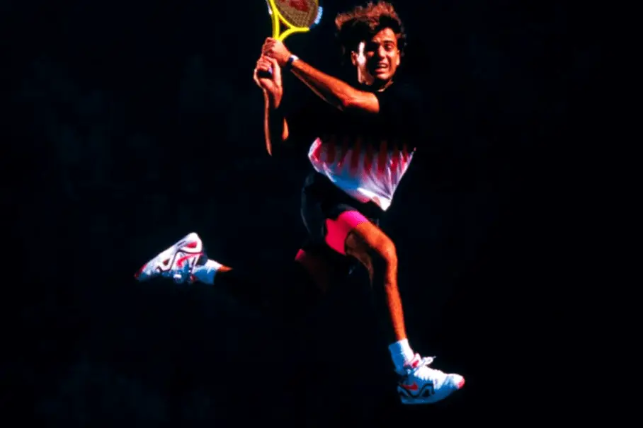 Andre Agassi leaping while holding a tennis racquet and wearing Nike Air Tech Challenge in front of a black background.