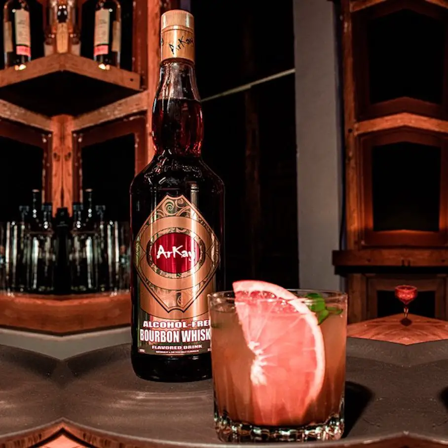 A bottle of ArKay Non-alcoholic Bourbon sitting next to a crystal cocktail glass.