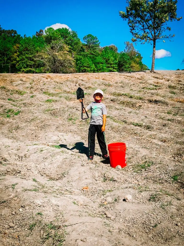 A young boy wearing a baseball cap stands in the middle of a field, holding a shovel in his hand. The field is at Crater of Diamonds State Park.