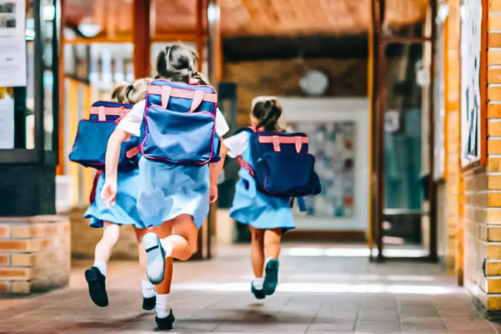 Three girls in blue dresses and backpacks running down a brick hallway.