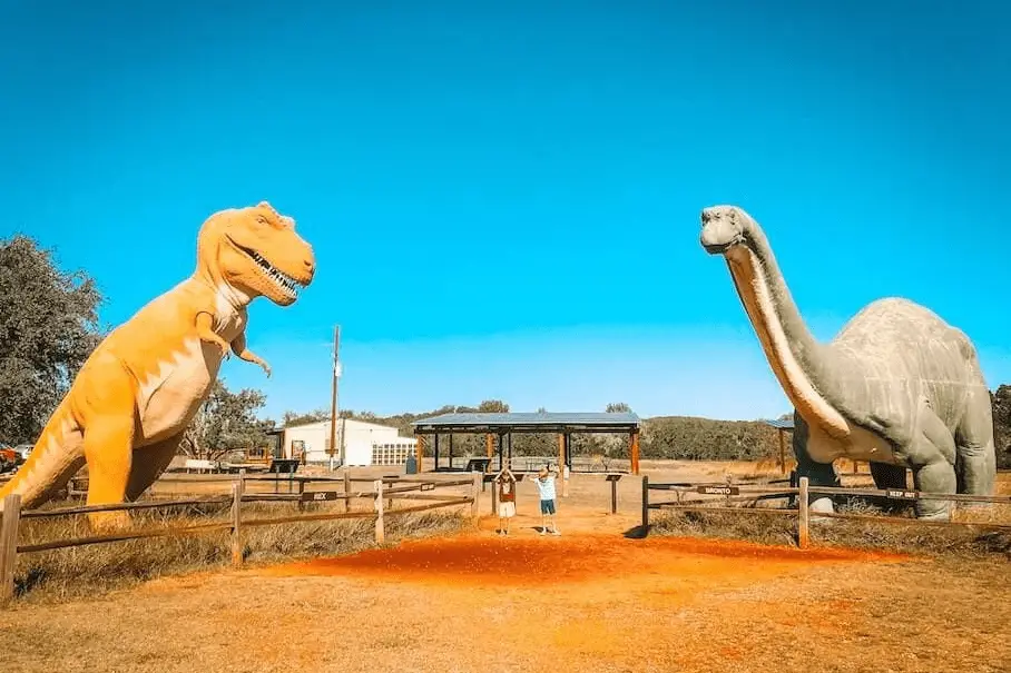 A photo of two dinosaur statues, a yellow T-Rex and a gray Brachiosaurus, in a park with two kids in the background.