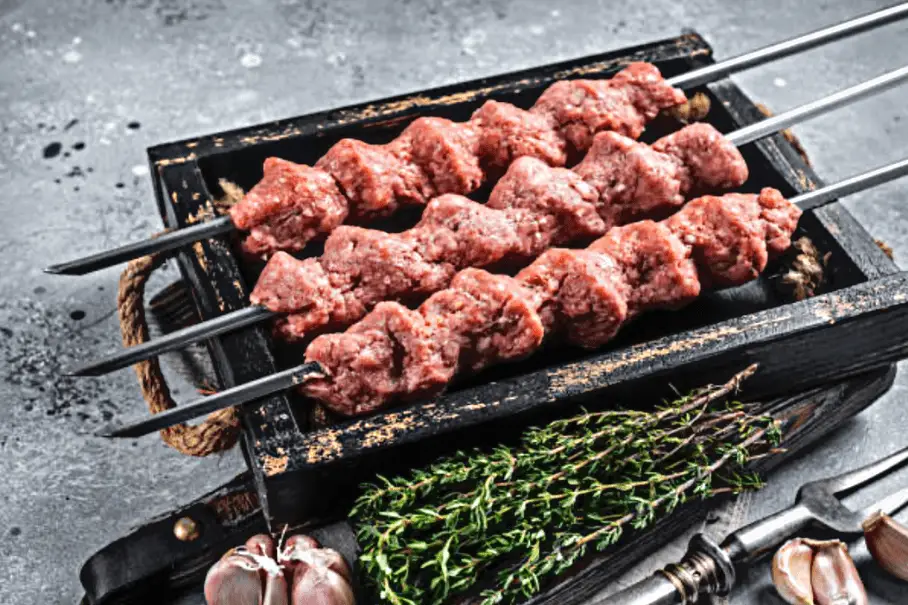 A photo realistic image of a grill pan with skewers of raw pork and some seasonings on a gray countertop.