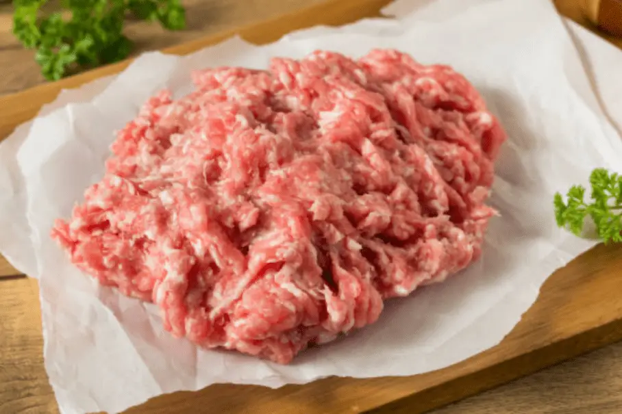 A pile of raw ground meat on parchment paper on a wooden cutting board with parsley.