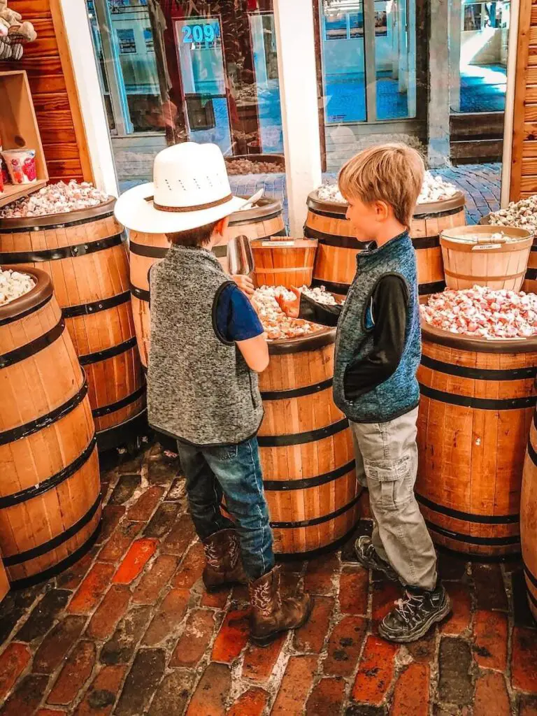 Two children standing in front of wooden barrels of candy in a store, with one of them wearing a cowboy hat. The store has a brick floor and a wooden exterior, and a window with a sale sign and a street outside.