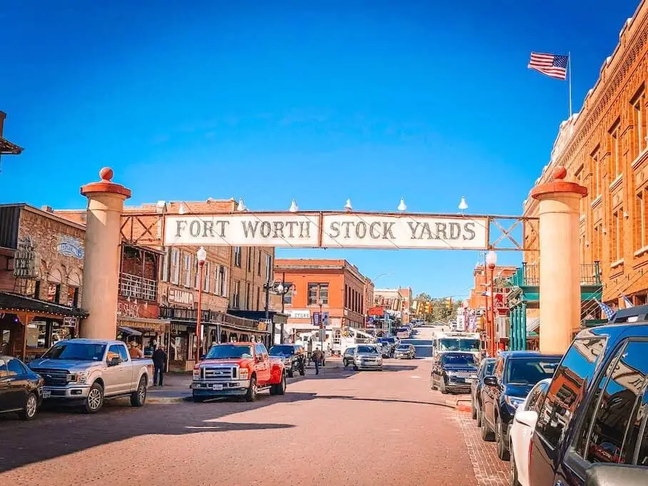 A street in Fort Worth Stock Yards, Texas, with cars parked on both sides and brick buildings with a western style. There is a large archway over the street that says “Fort Worth Stock Yards” and an American flag on the right side. The sky is blue and the street is busy with people and cars.