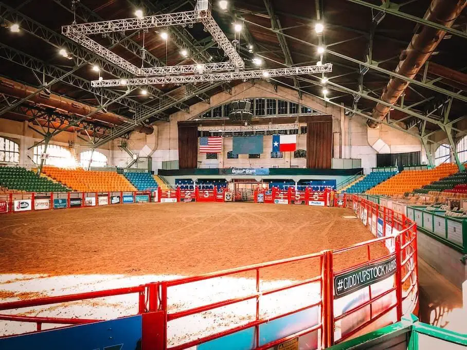 A photo realistic image of an empty indoor rodeo arena with a dirt floor, colorful seats, flags and banners at the Fort Worth Stockyards.