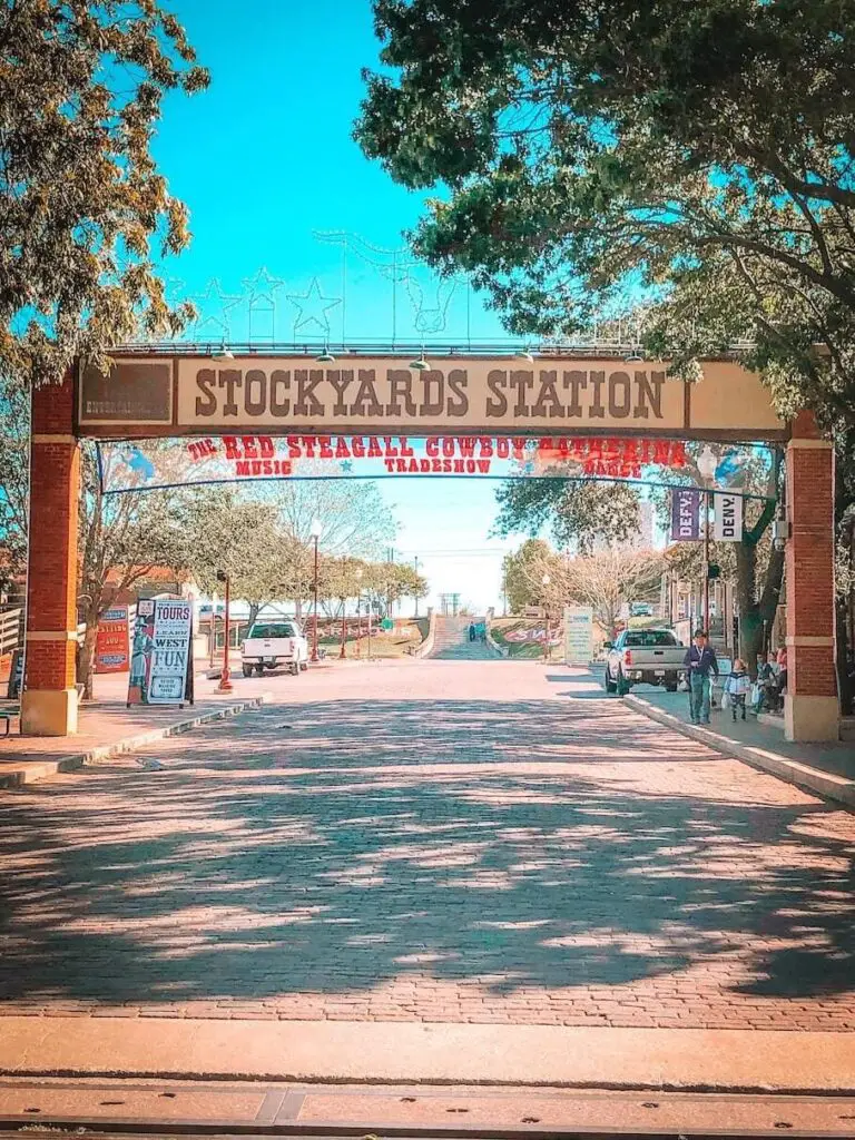 The entrance to Stockyards Station in Fort Worth, Texas, with a metal arch that says “Stockyards Station” in red letters, and two brick pillars that say “The Texas Cowboy Hall of Fame” and “Billy Bob’s Texas”. The entrance is on a brick street with trees on both sides and a blue sky
