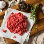 A photo realistic image of raw minced meat on a wooden cutting board with salt, pepper and rosemary on a beige cloth background.