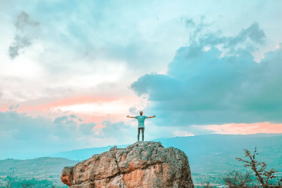 A person standing on a rock with their arms outstretched in front of a mountain landscape with a blue and pink sky