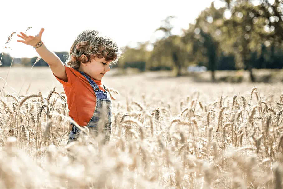 A kid in an orange shirt and blue overalls standing in a golden field of tall grass with their arms outstretched and looking away from the camera.