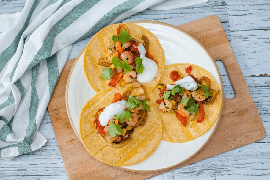 Three tacos with grilled shrimp, bell peppers, onions, cilantro, and sour cream on yellow corn tortillas, on a wooden cutting board on a white wooden table with a striped towel on the side.