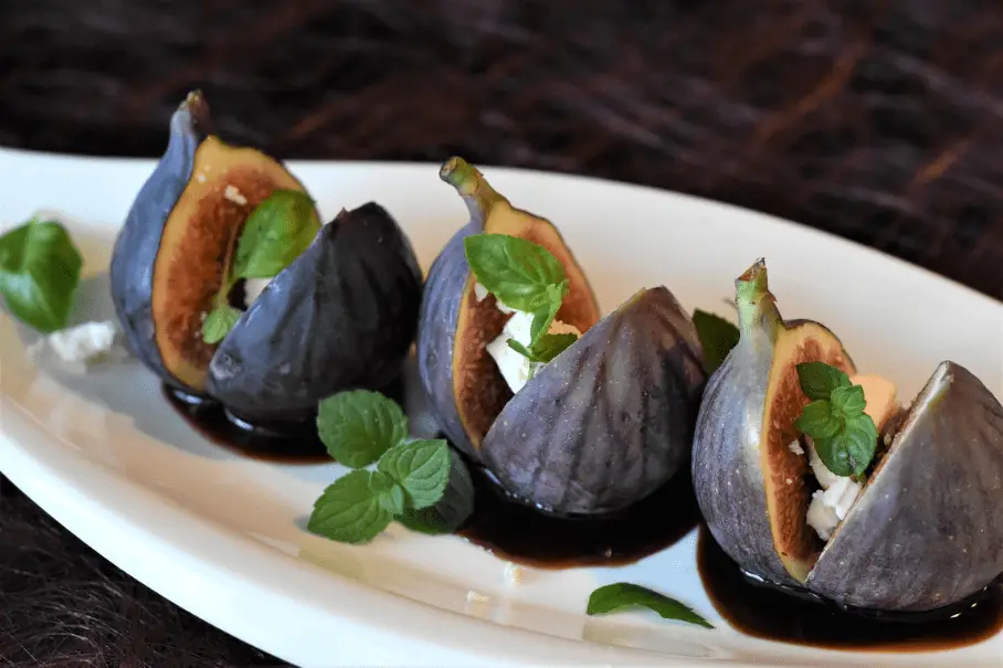 A white plate with five figs cut in half and drizzled with a dark sauce and mint leaves, on a dark wooden table. The background is blurred and not in focus.