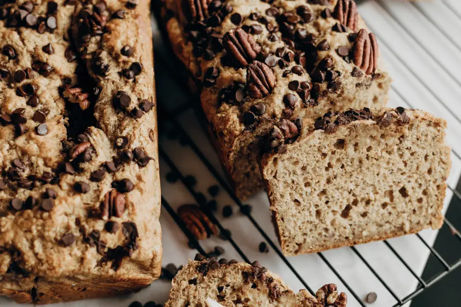 Two loaves of banana bread with pecans and chocolate chips on a wire cooling rack, with one loaf sliced open. The bread is freshly baked and on a white countertop. The background is a blurred kitchen setting.