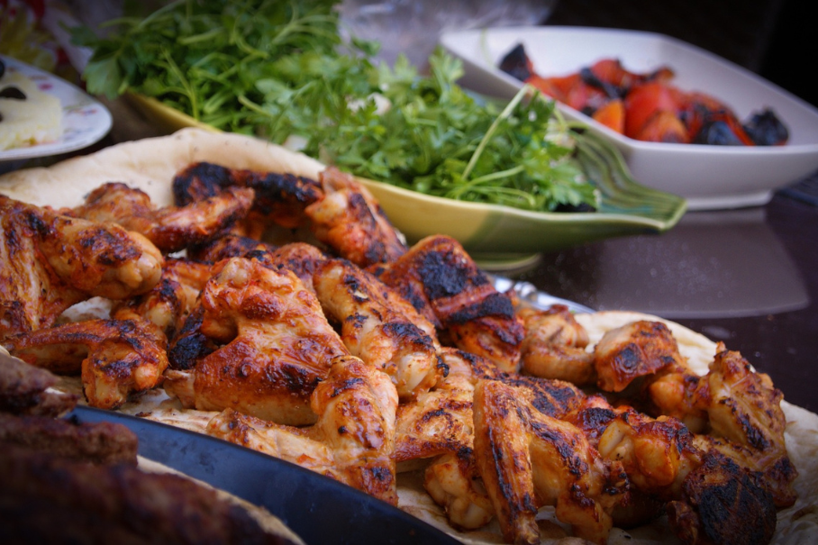 A platter of grilled chicken wings, with a green salad and red grilled peppers on white plates in the background. The background is a blurred table with other dishes on it.