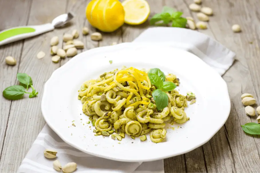 A plate of yellow pasta with basil and pistachio on a wooden table, with a lemon and a spoon on the side. The plate is white and the background is a blurred kitchen or dining area.