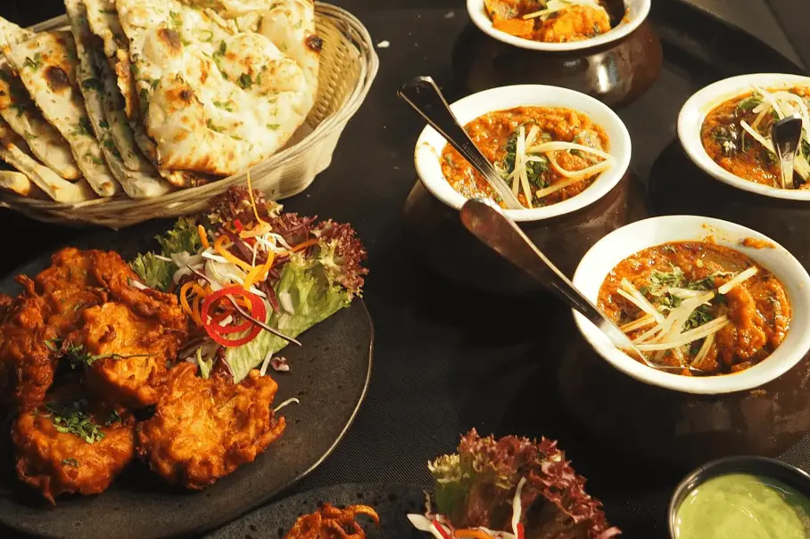 A photo realistic image of an Indian meal with a bowl of chicken curry, various condiments, and breads on a brown tablecloth.