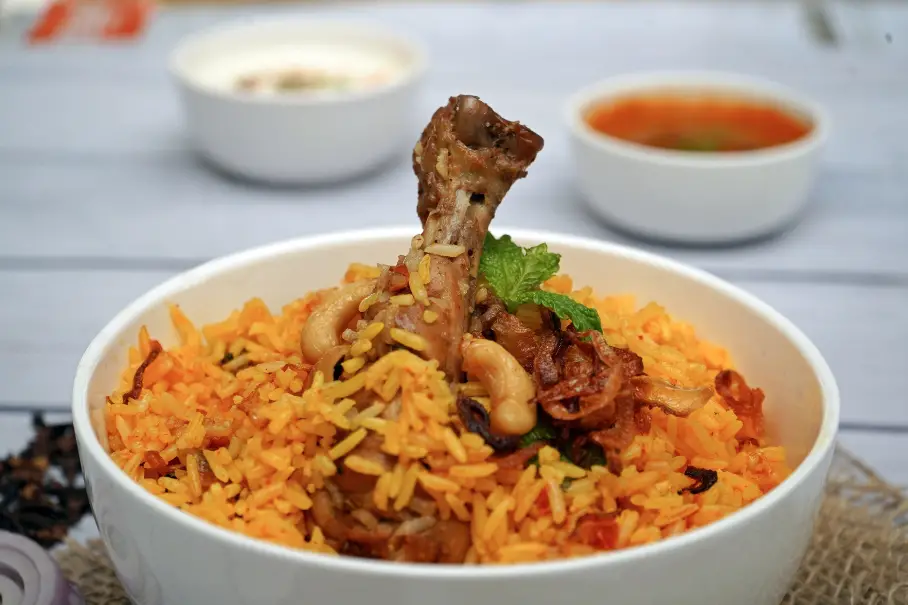 A bowl of biryani with a lamb shank on top, served on a white wooden table with two bowls of sauce and a plate of black sesame seeds. The biryani has orange and yellow rice with meat and vegetables, and a mint leaf on top. The lamb shank is brown and cooked.