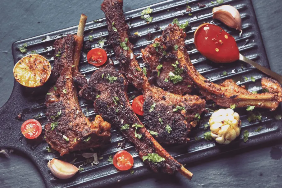 A black grill pan with grilled lamb chops, cherry tomatoes, garlic, and lemon on a dark gray surface, with a wooden spoon on the edge of the pan. The lamb chops are spiced and herb-garnished.