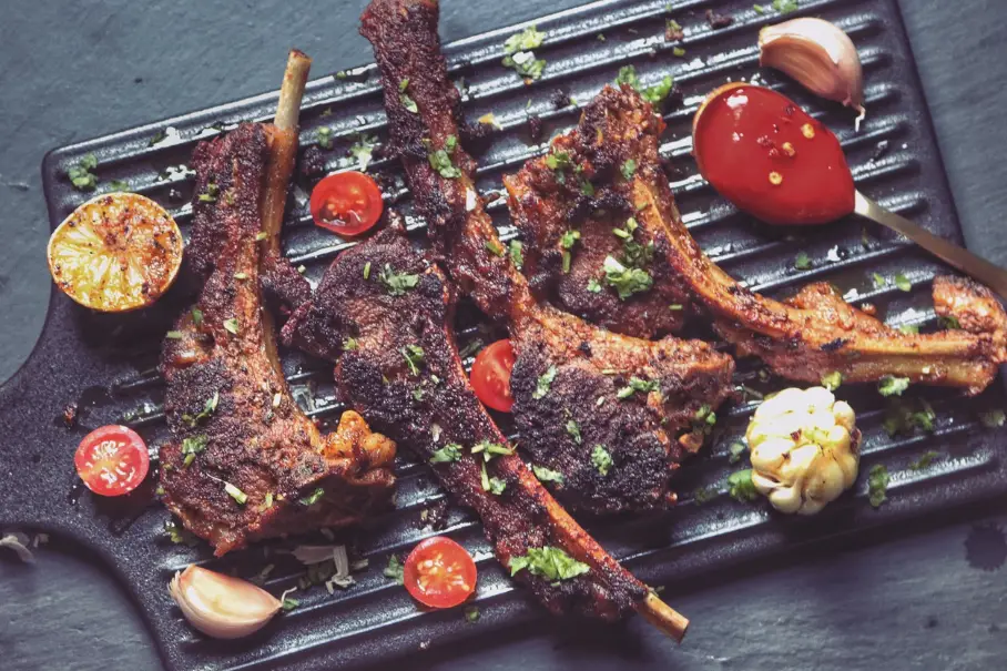 A black grill pan with grilled lamb chops, cherry tomatoes, garlic, and lemon on a dark gray surface, with a wooden spoon on the edge of the pan. The lamb chops are spiced and herb-garnished.