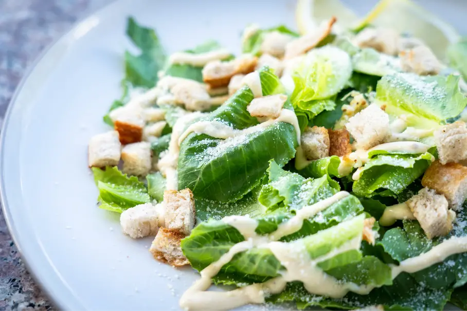 A photo realistic image of a Caesar salad on a white plate. The salad has romaine lettuce, croutons, Parmesan cheese, and a creamy dressing. The plate is on a gray countertop with a blurred background.