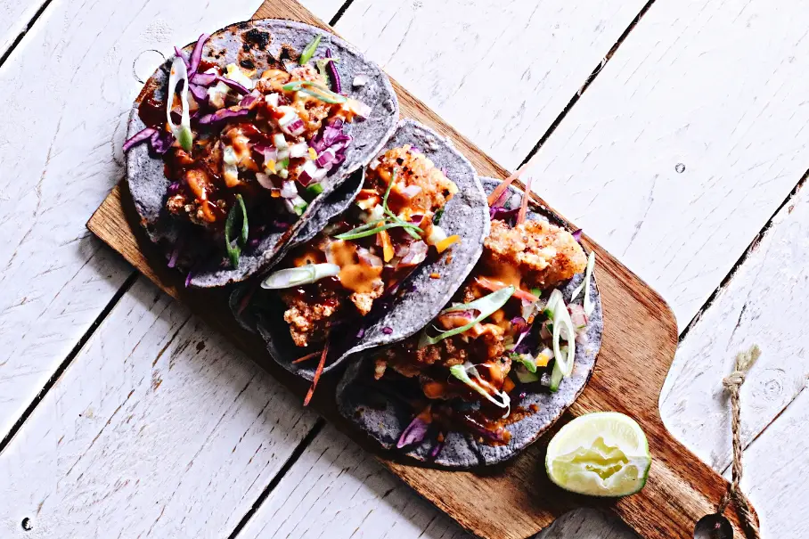 A photo realistic image of three blue corn tacos with fried fish, red cabbage, and a creamy sauce on a wooden serving board. The board has a handle and is placed on a white wooden table with a lime wedge and a cilantro sprig. The image has a bright and fresh mood.