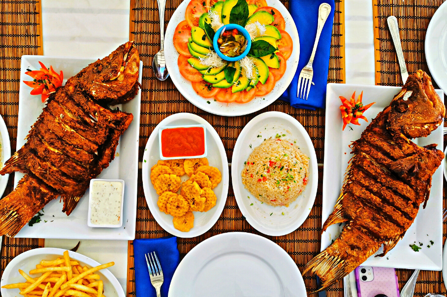 A photo of a table with a seafood feast on a checkered tablecloth. The table has two grilled fish, a plate of shrimp, fried calamari, rice, french fries, and two sauces.