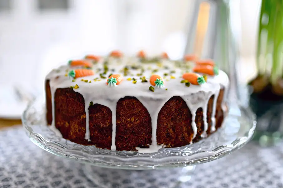 A carrot cake with white icing and carrot decorations on a glass cake stand.