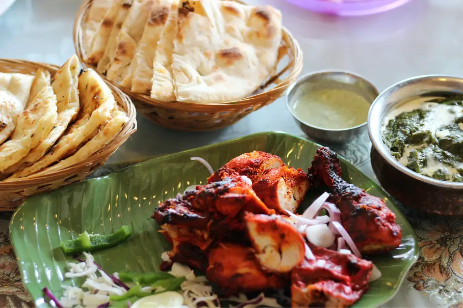 A photo of an Indian meal with tandoori chicken, naan bread, chutney, and palak paneer on a gold tablecloth.