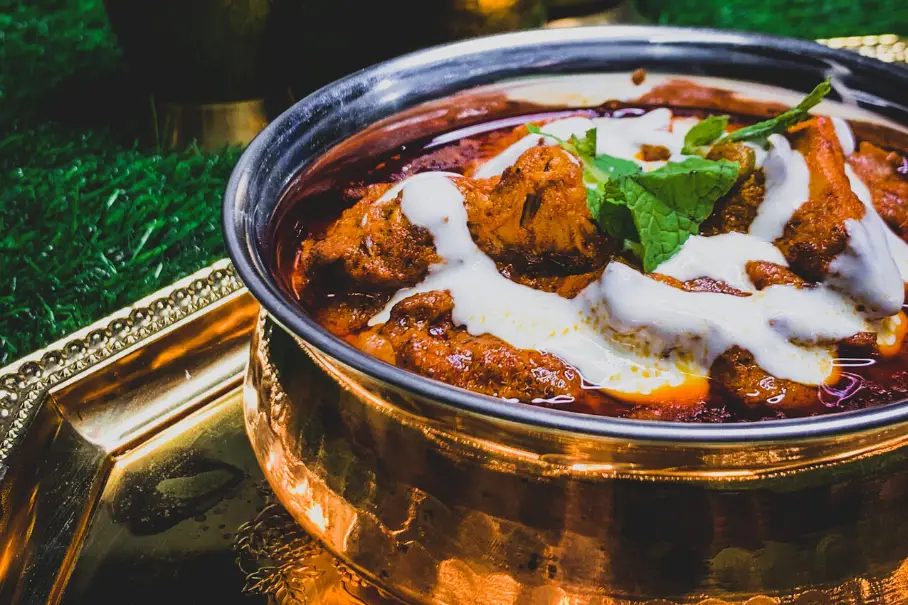 A photo of a dish of Indian chicken tikka masala with meat and sauce in a copper dish on a gold tray on a green mat.