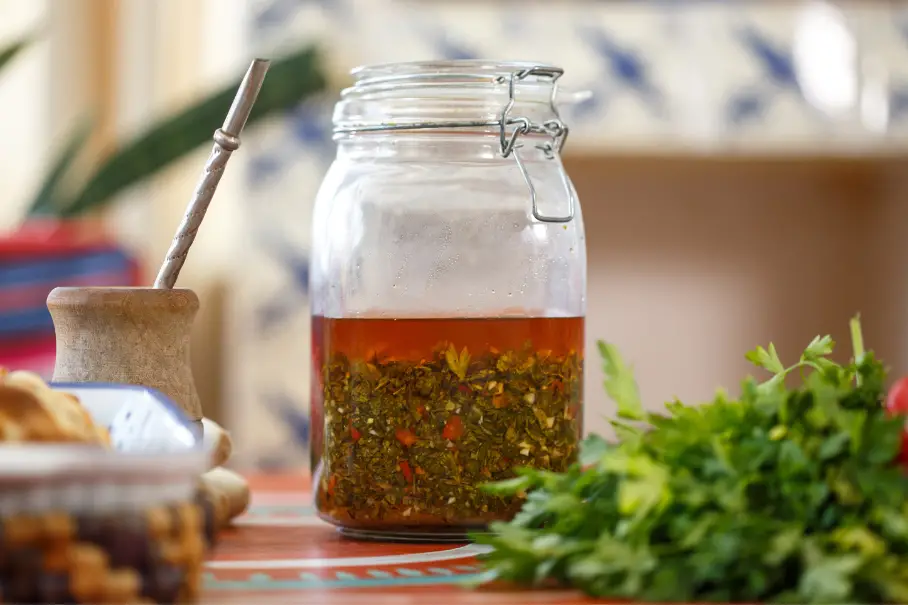 A photo of a jar of chimichurri with herbs and a wooden spoon on a kitchen table.