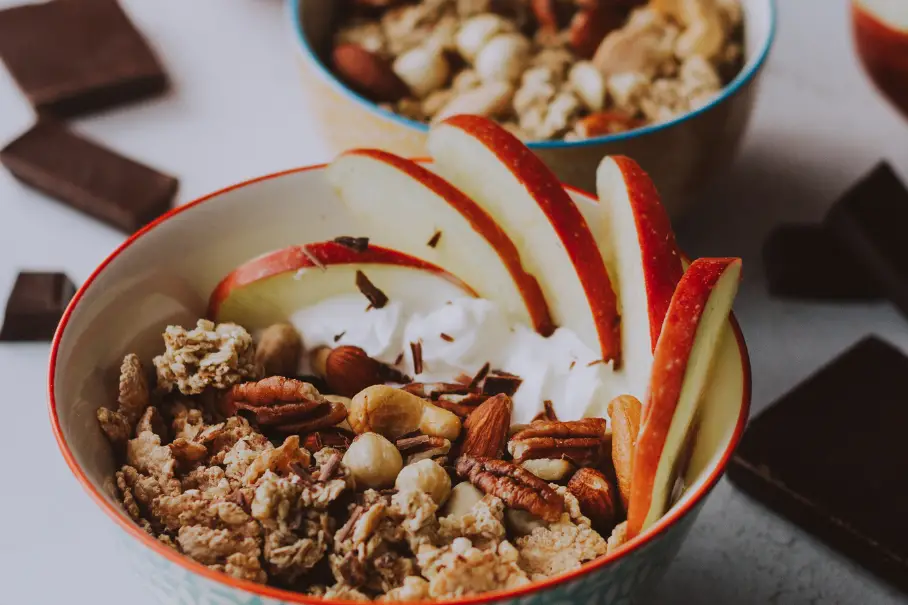Two bowls of granola with sliced apples and nuts on top, on a white countertop with chocolate bars around. The bowls are blue and red, the granola is golden brown, the apples are thin and fanned, and the nuts are pecans and hazelnuts.