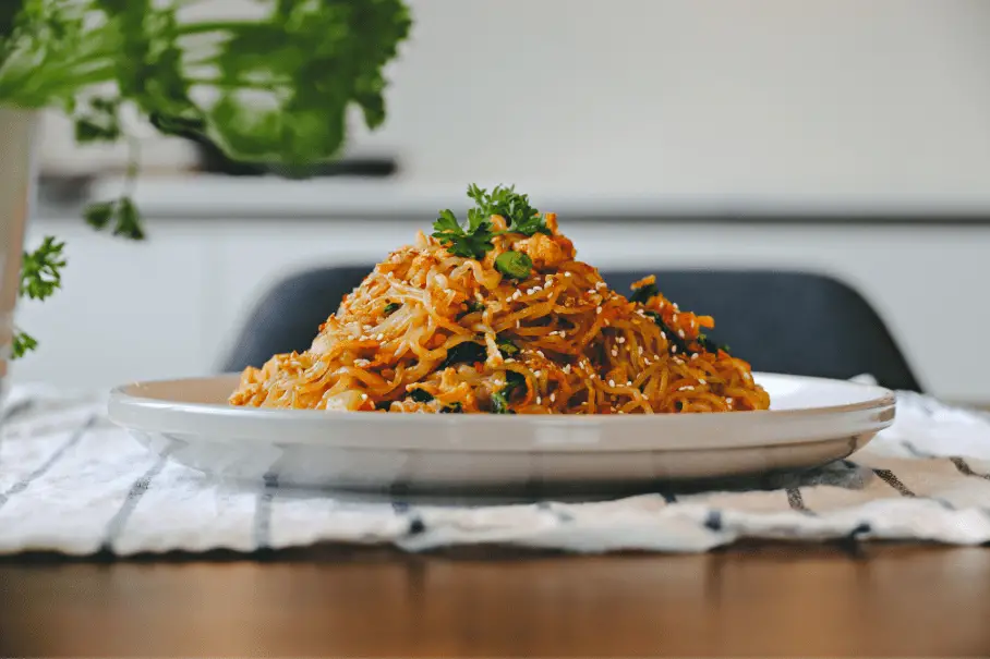 A plate of Pad Thai noodles with herbs on a checkered tablecloth with a chair and a plant in the background.