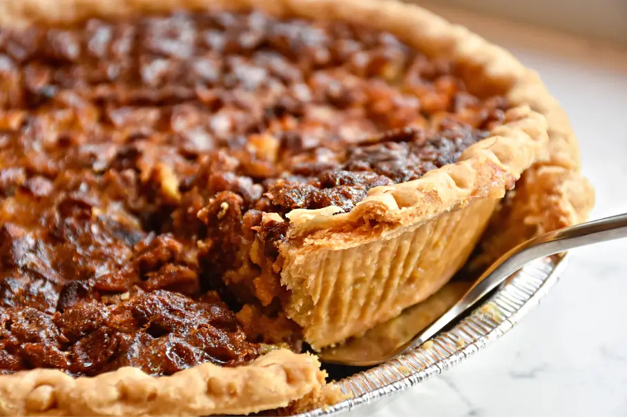 A photo realistic image of a pecan pie with a slice being taken out of it. The pie has a golden brown crust and a dark brown filling with pecans on top. The slice being taken out of the pie is on a silver pie server. The pie is on a white marble countertop. The background is blurred and not in focus.
