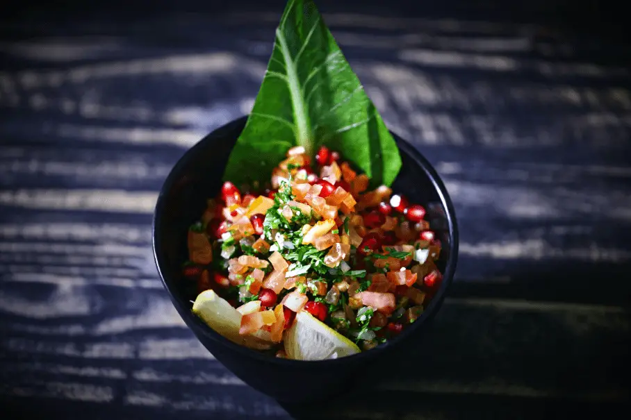 An image of a black bowl of pico de gallo on a black background. The pico de gallo is made of diced tomatoes, onions, cilantro, and other spices. It is garnished with a large lettuce leaf.